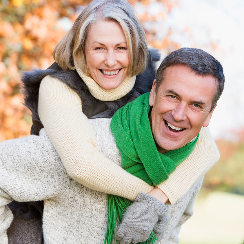 An outdoor scene where a man wearing a green scarf and sweater gives a piggyback ride to a woman dressed in a fur coat and sweater, both smiling happily.
