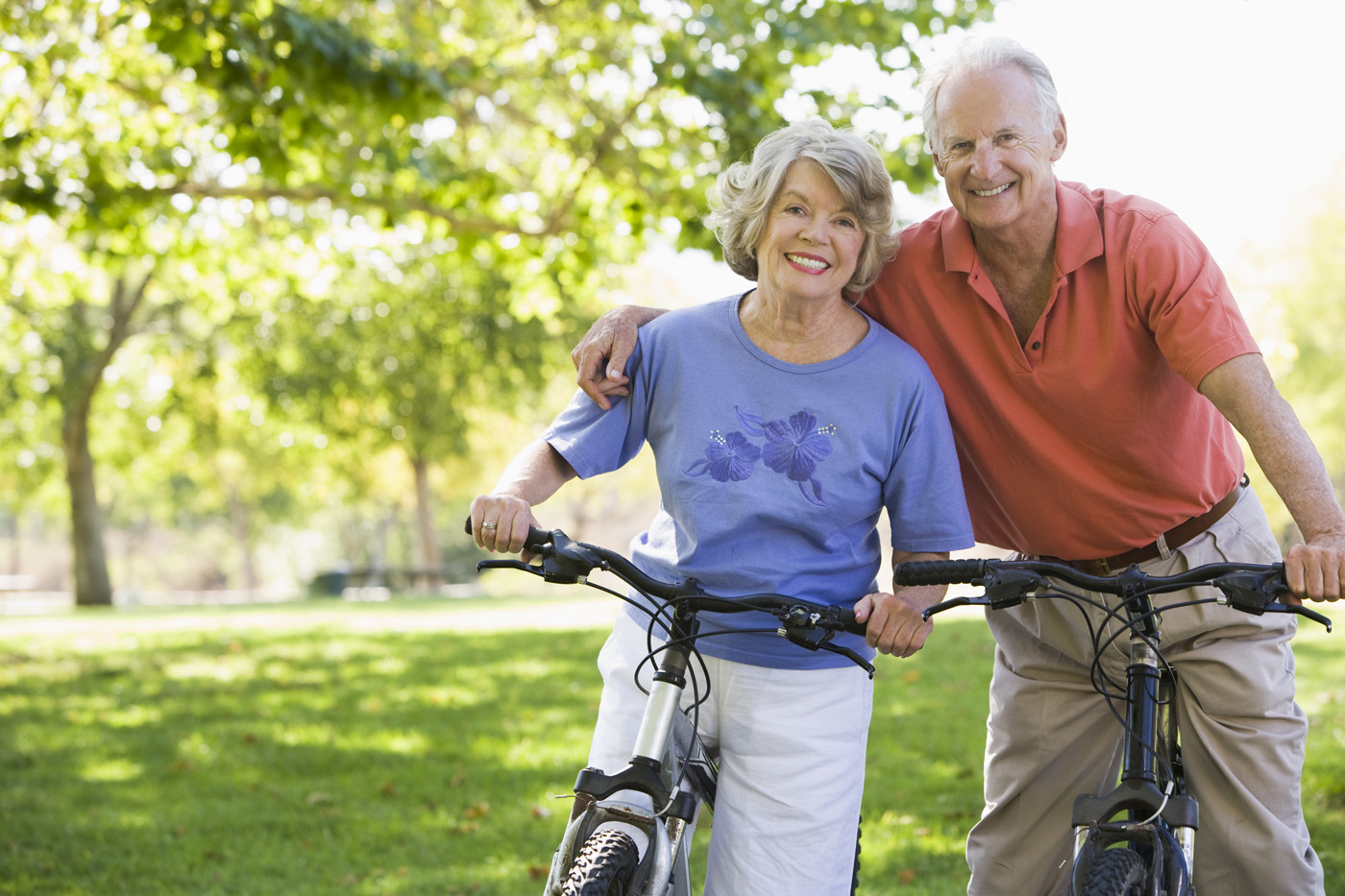 The elderly couple both riding bicycles, with the smiling man in the front wrapping his arm around the woman behind.