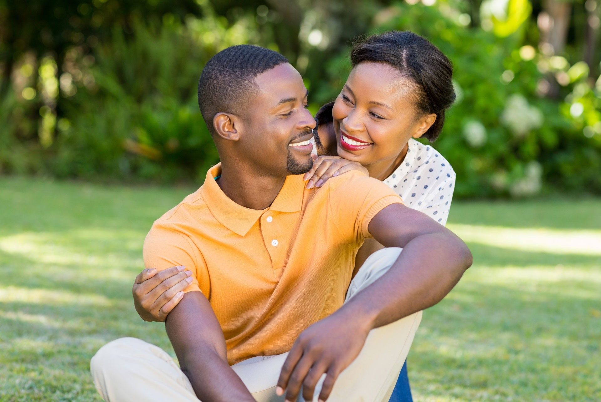 A smiling man and woman, appearing as a couple, showcase their healthy teeth while in an outdoor setting surrounded by lush green nature.