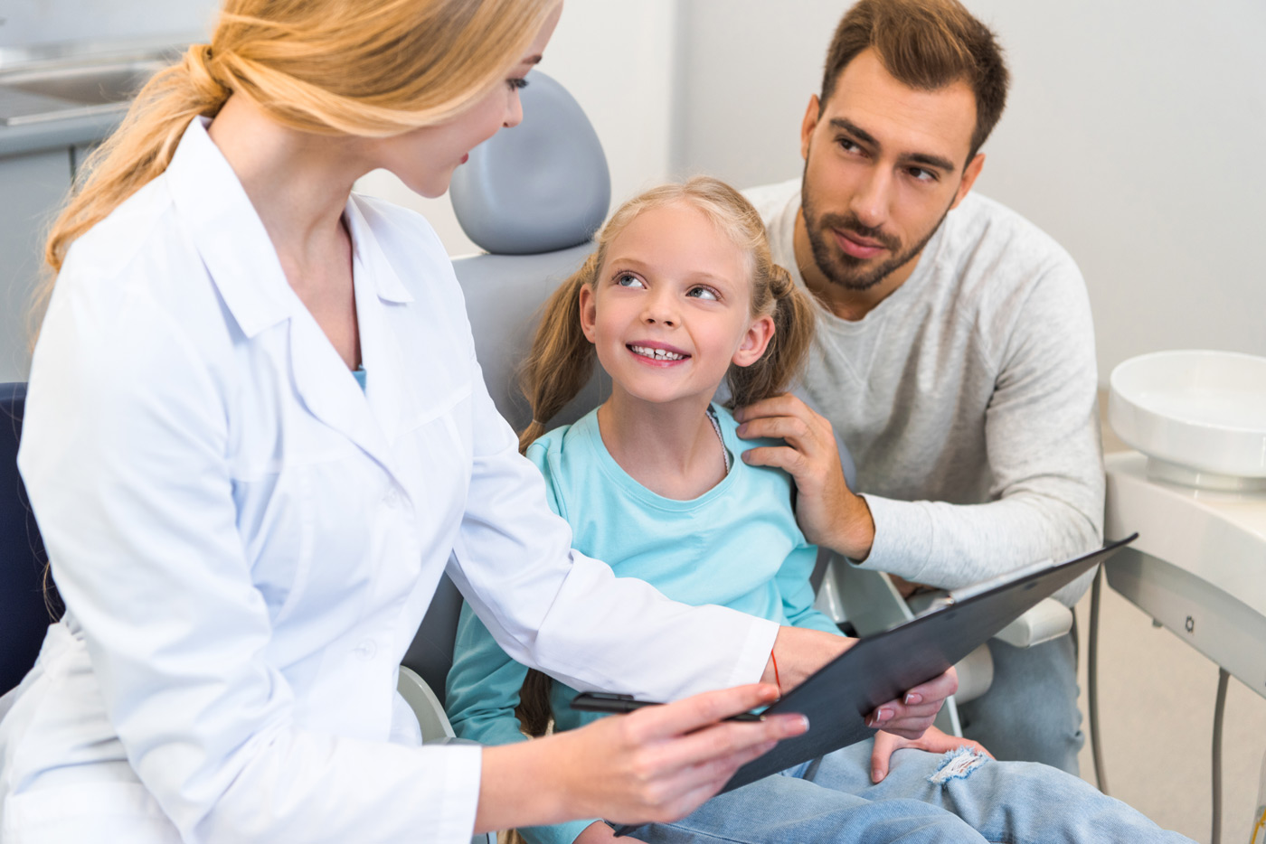 In an orthodontic consultation, a dentist showing a dental planner a child in the chair, while a comforting father looks on.