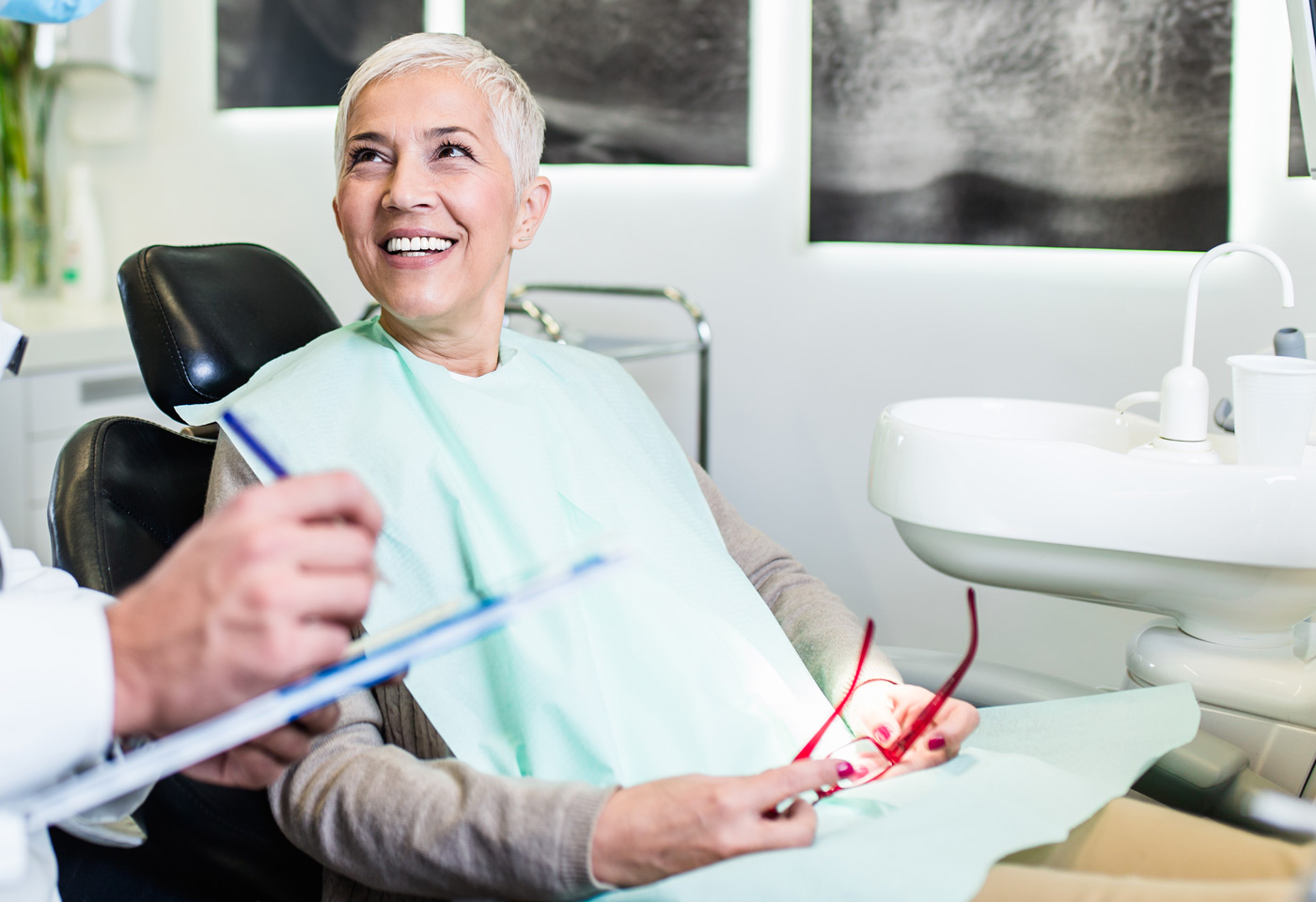 A woman sitting in a dental chair, smiling and holding her eyeglasses, looks towards a dentist who is holding a pen and clipboard, writing or discussing a dental procedure.
